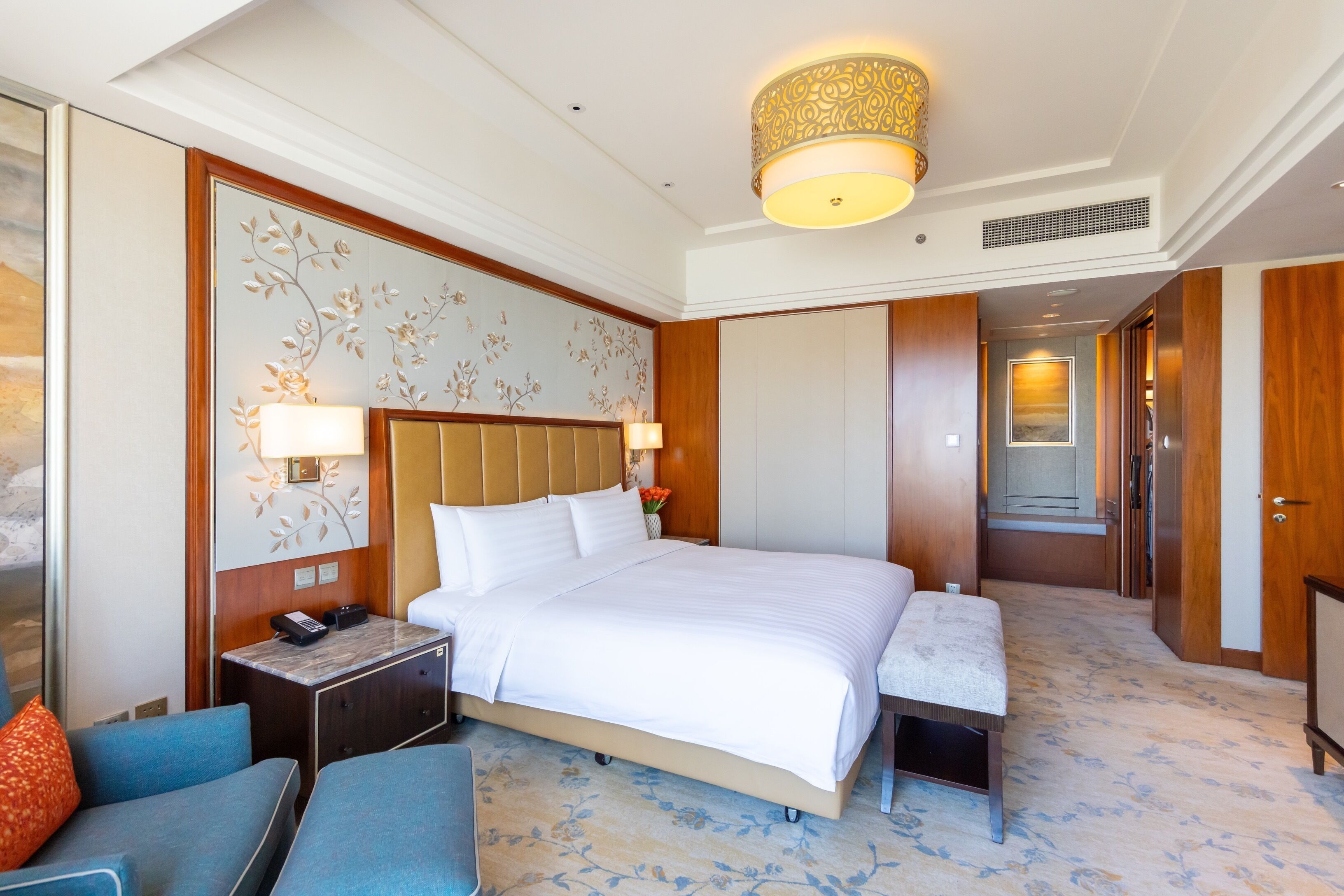 A hotel room with a large bed, a bedside table with a lamp, a small desk, a chair, and a bench.