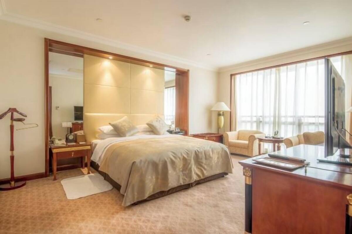 Quarto de hotel com uma cama grande, uma mesa, uma cadeira, uma televisão e uma janela ampla.