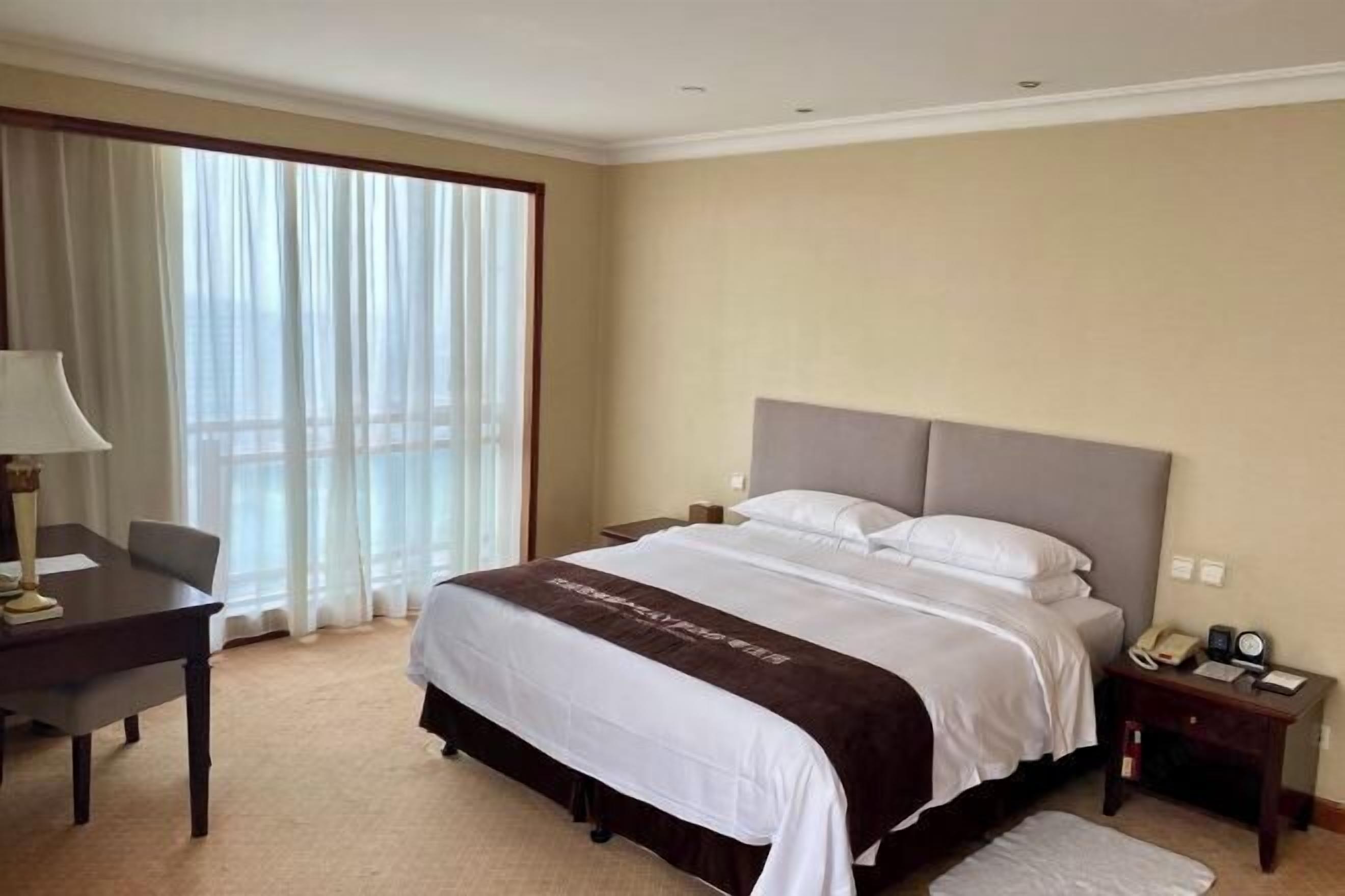 A hotel room with a large bed, a desk, a chair, a nightstand, and a window with curtains.
