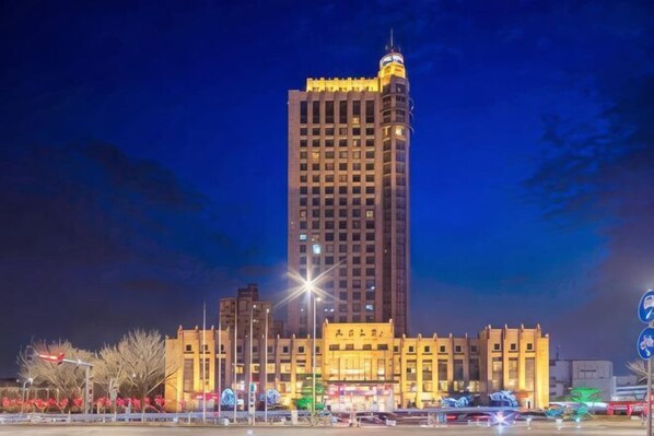 Un edificio alto e illuminato con una torre d'angolo ben visibile, circondato da una strada trafficata con veicoli e lampioni.
