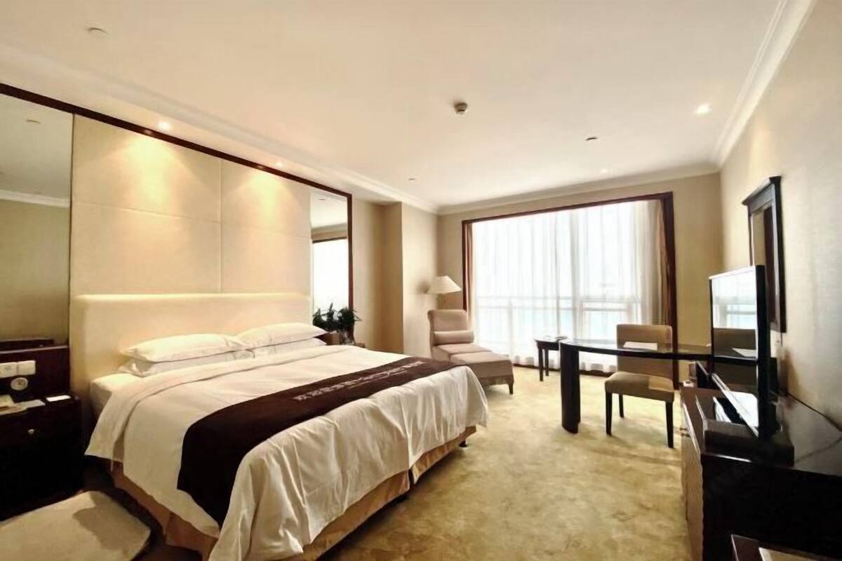 Quarto de hotel com cama grande, escrivaninha, cadeira, televisão e janela ampla.