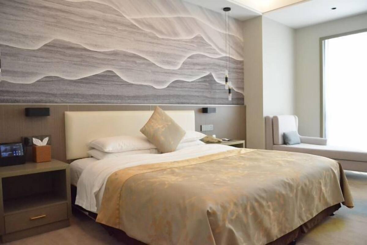 Quarto de hotel com cama, mesa de cabeceira, luminária na parede e janela com vista.