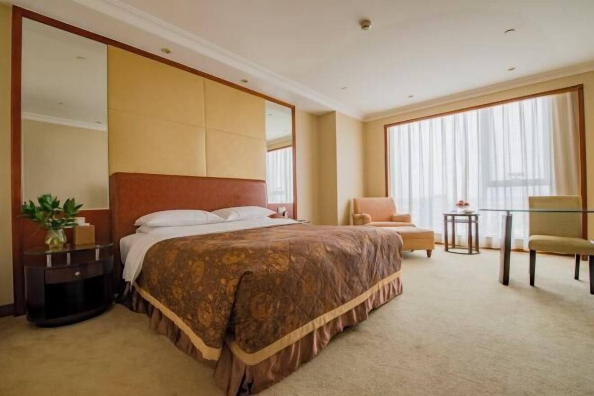 Quarto de hotel com uma cama grande, uma cadeira, uma mesinha e uma janela ampla com cortinas.