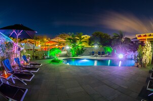Un área de piscina exterior de noche, con sillones, sombrillas y una pared de piedra.