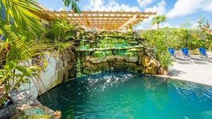 Un área de piscina con una cascada, rodeada de plantas tropicales y una pérgola de madera.