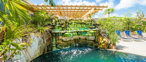 A pool area with a waterfall feature, surrounded by tropical plants and a wooden pergola.