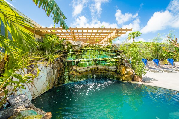 A pool area with a waterfall feature, surrounded by tropical plants and a wooden pergola.