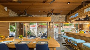 Un restaurante con interior de madera, una zona de bar y mesas al aire libre visibles a través de amplios ventanales.