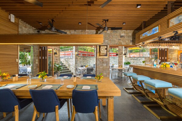 Un restaurante con interior de madera, una zona de bar y mesas al aire libre visibles a través de amplios ventanales.