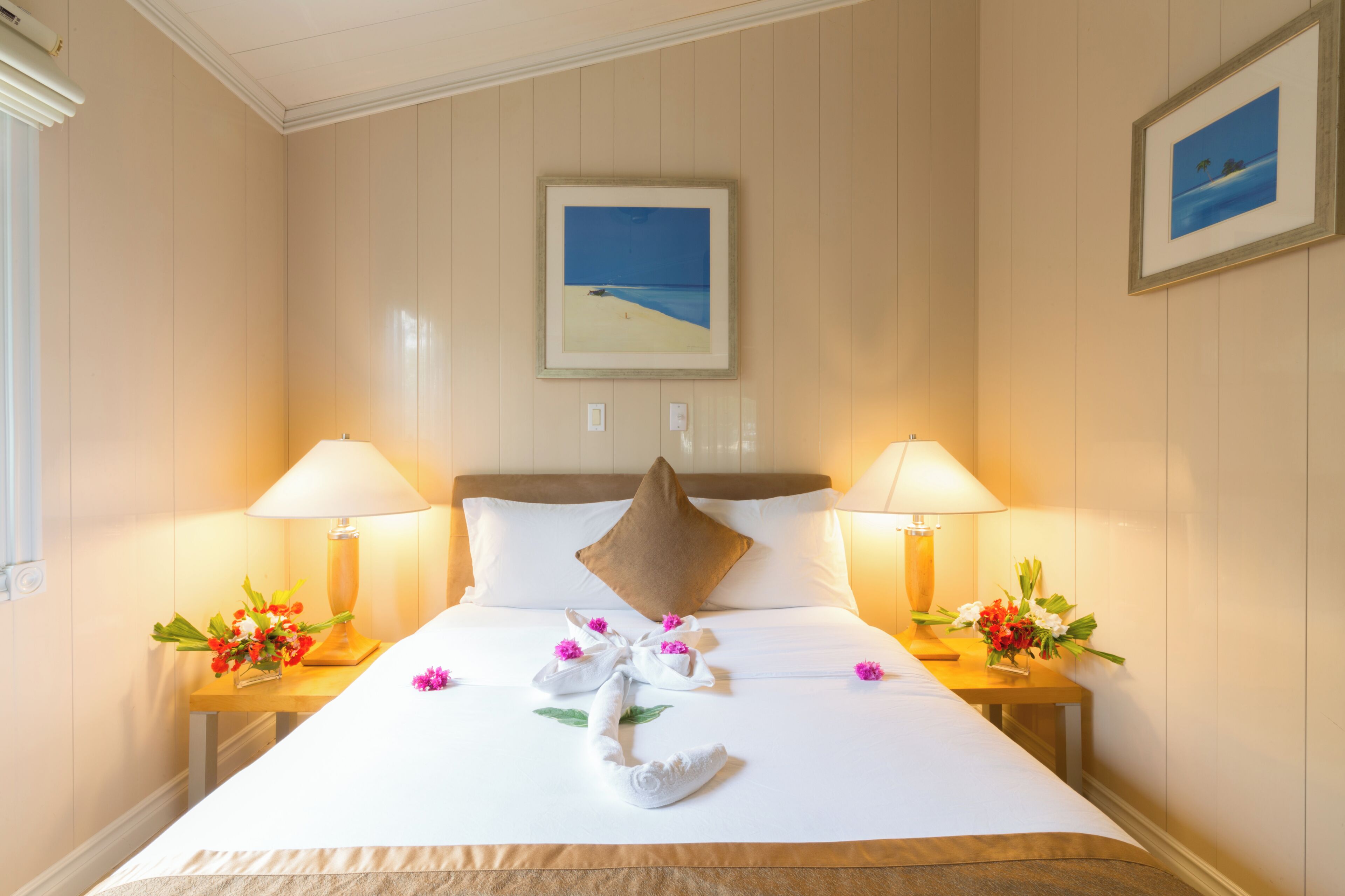 Una cama bien hecha con un almohadón, dos lámparas de noche y arreglos florales sobre mesitas de madera.