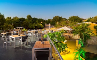 An outdoor dining area with multiple tables and chairs, surrounded by greenery and a pool.