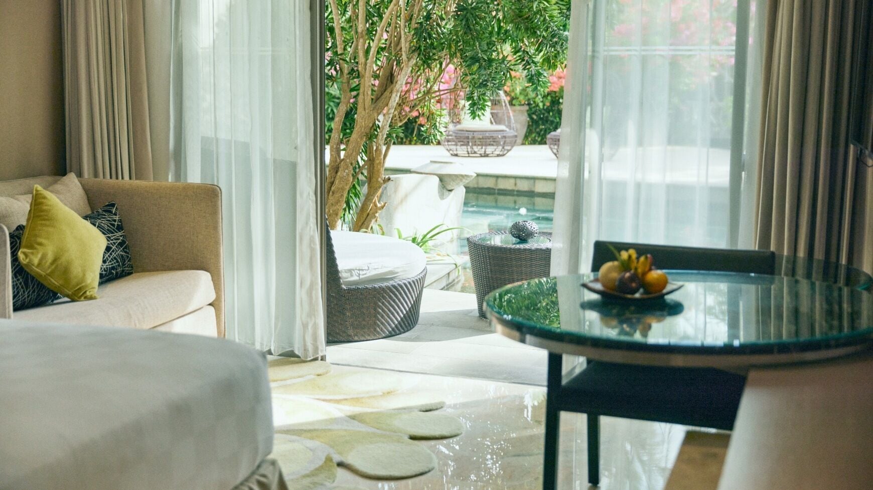 A modern living room with a sofa, a round glass table with fruit, and a view of a garden and pool.