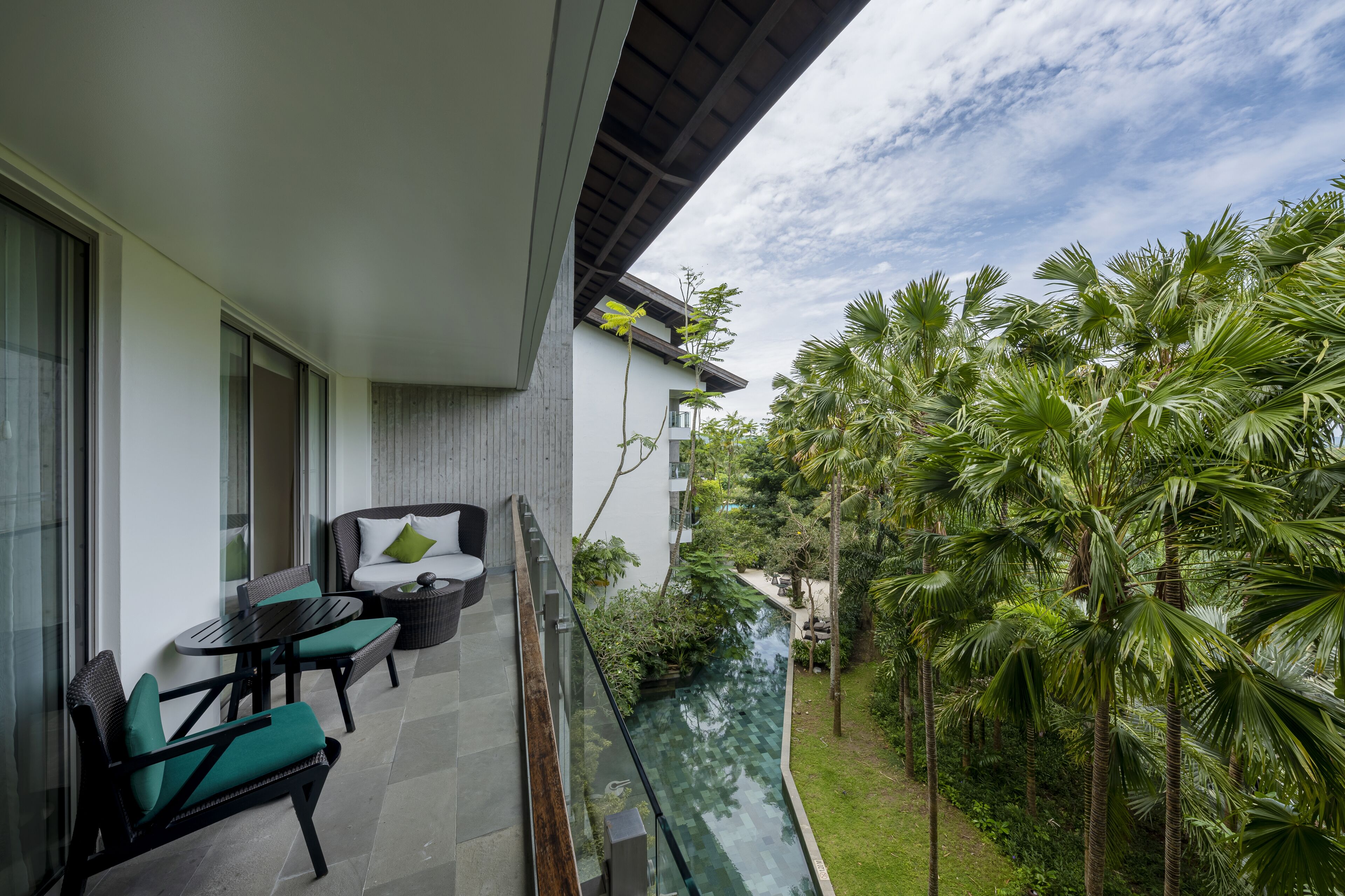 A modern balcony with a glass railing overlooking a lush garden and a pool.