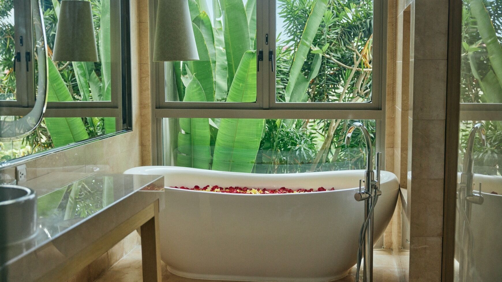 A modern bathroom with a freestanding bathtub filled with rose petals, large windows with a view of lush greenery, and a sleek, minimalist design.