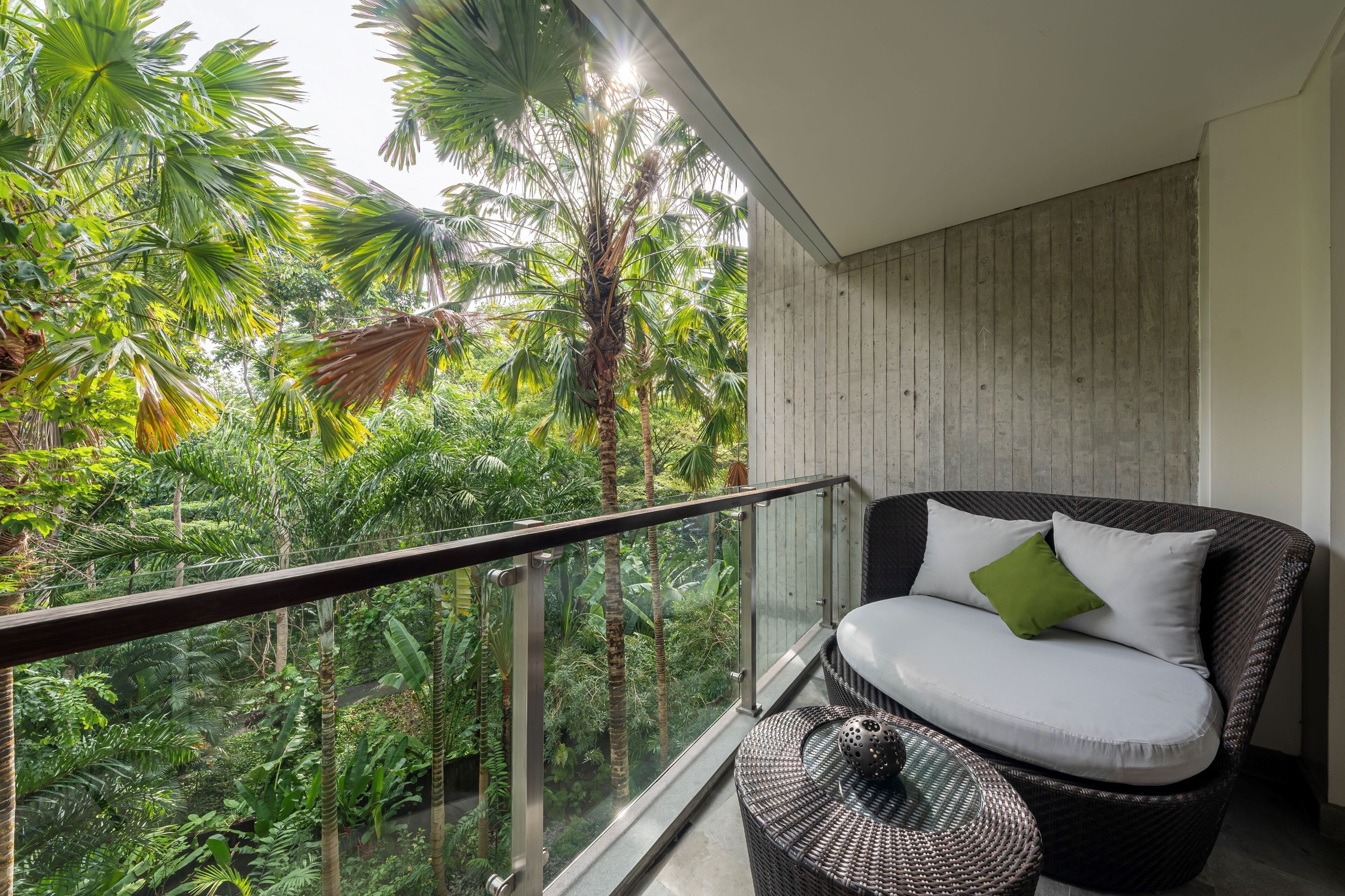 A balcony with a wicker chair and a small round table, overlooking a lush garden with various plants and trees.