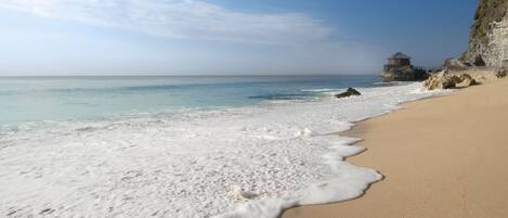 A beach with clear water, a sandy shore, and a rocky cliff with a structure on top.
