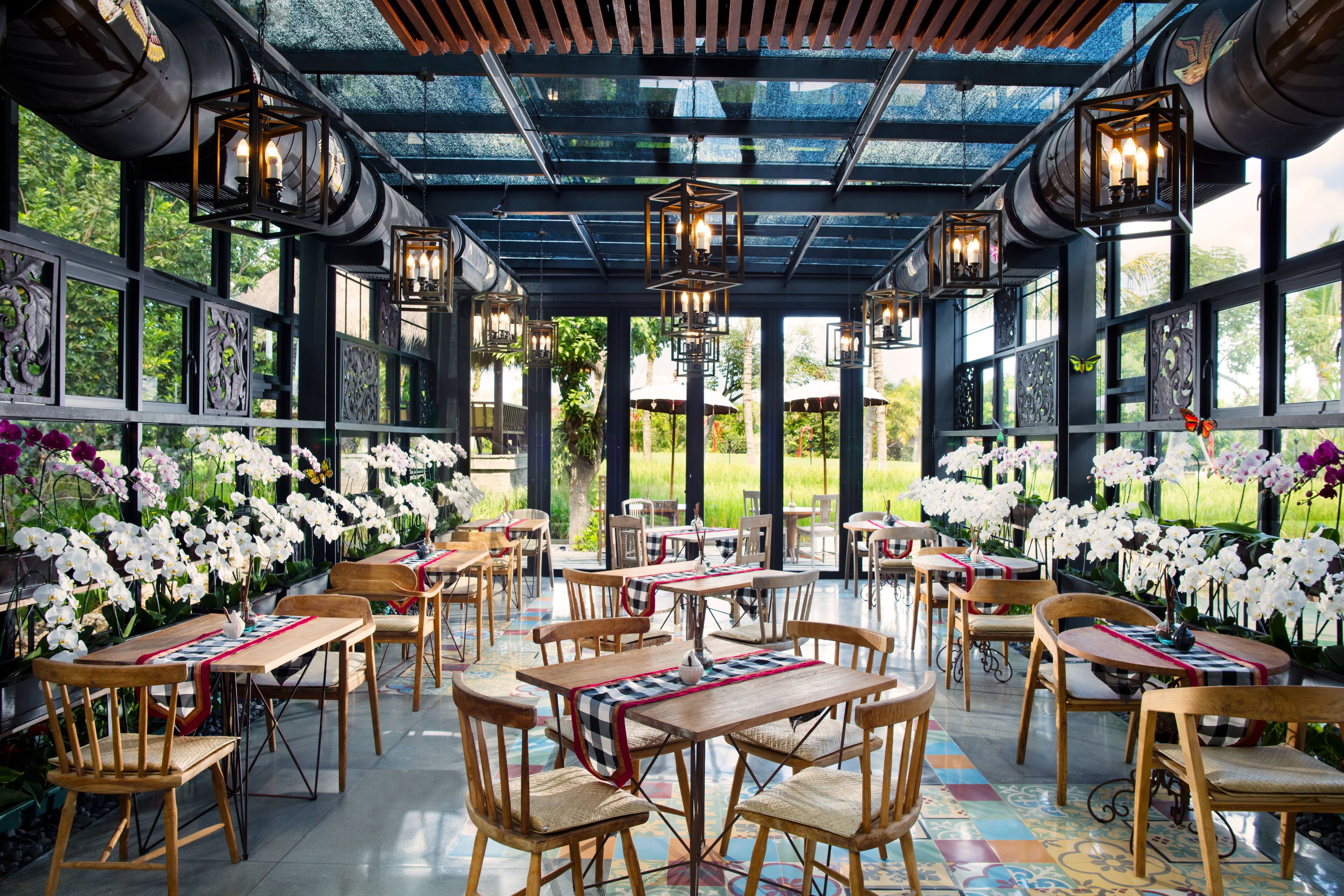An outdoor dining area with wooden tables and chairs, surrounded by white orchids and a glass ceiling.