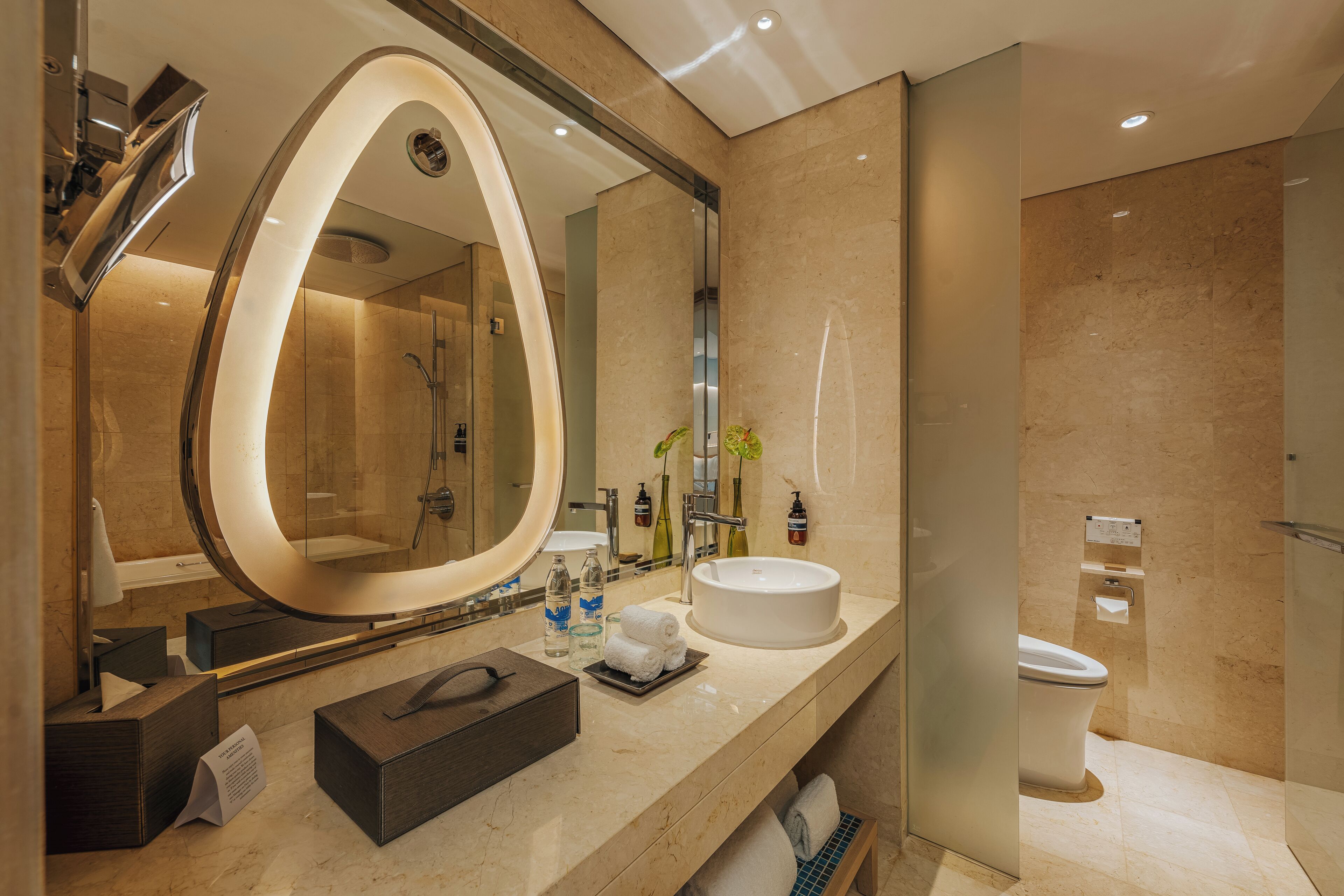 A modern bathroom with a large oval mirror, a white sink, and a glass-enclosed shower.