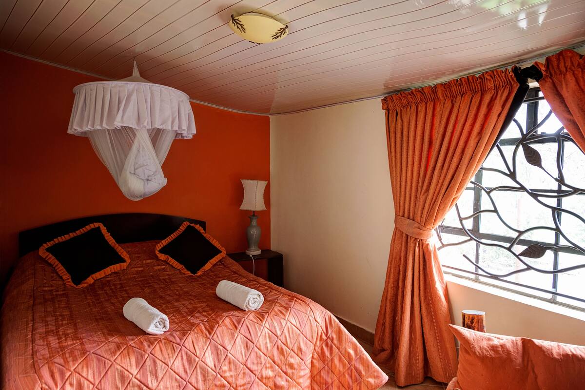 A neatly made bed with a quilted orange bedspread, two pillows, and two rolled towels. A white canopy hangs above the bed.
