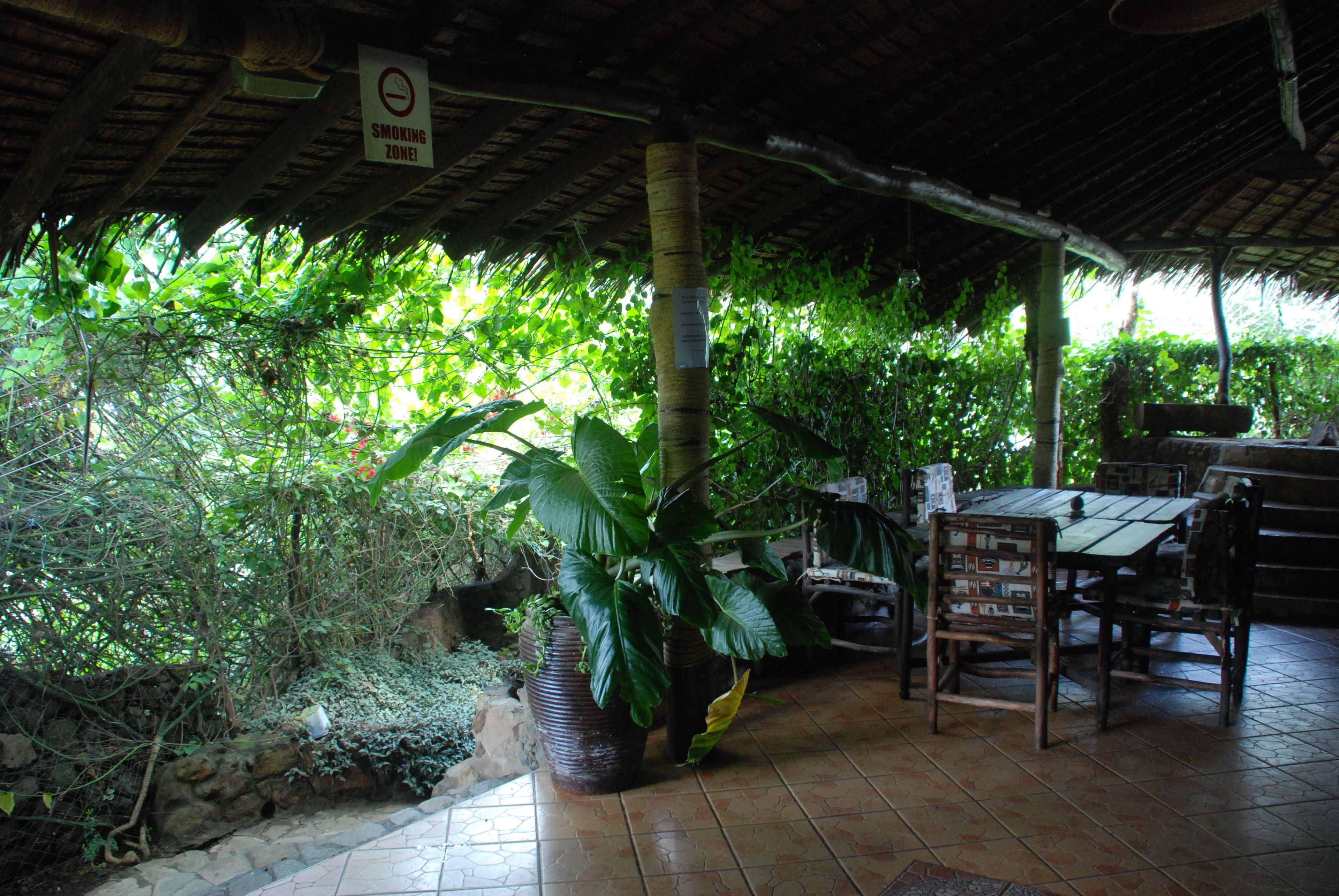 An outdoor covered patio with a smoking zone sign, surrounded by lush greenery and a tiled floor.
