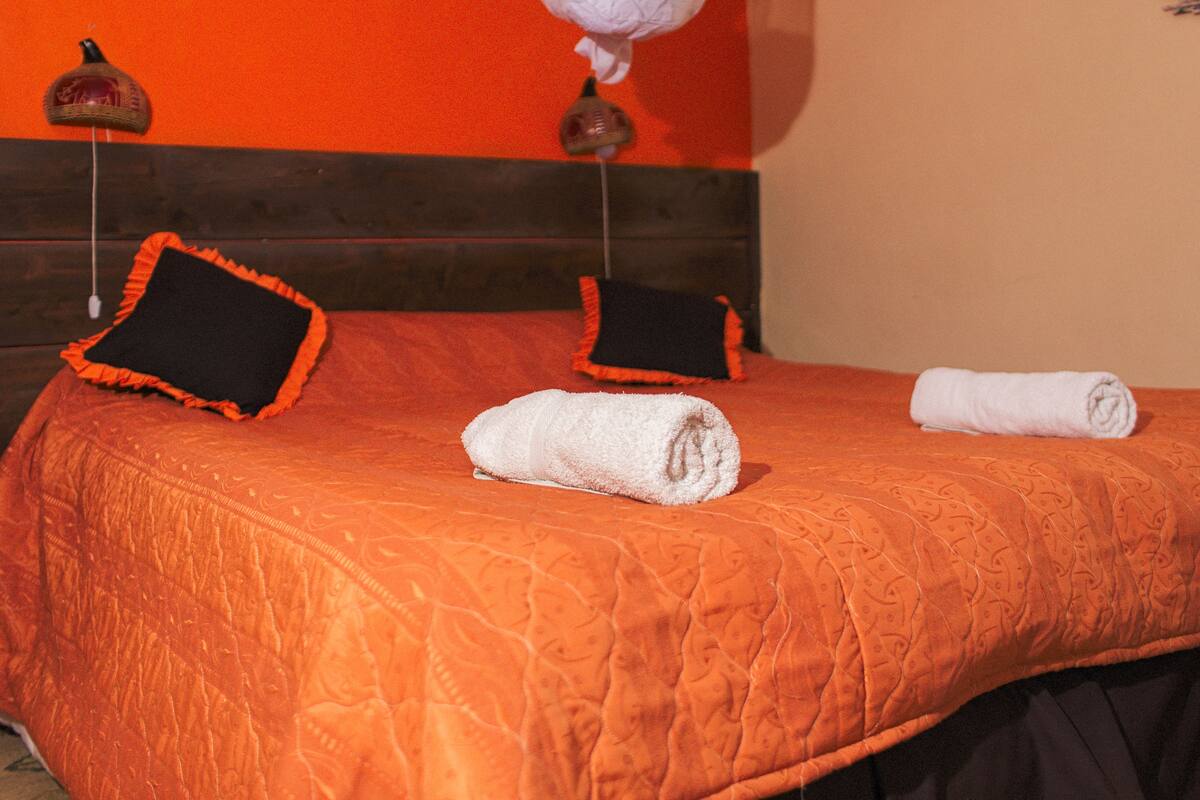 A bed with an orange quilt, two pillows, and a rolled towel.