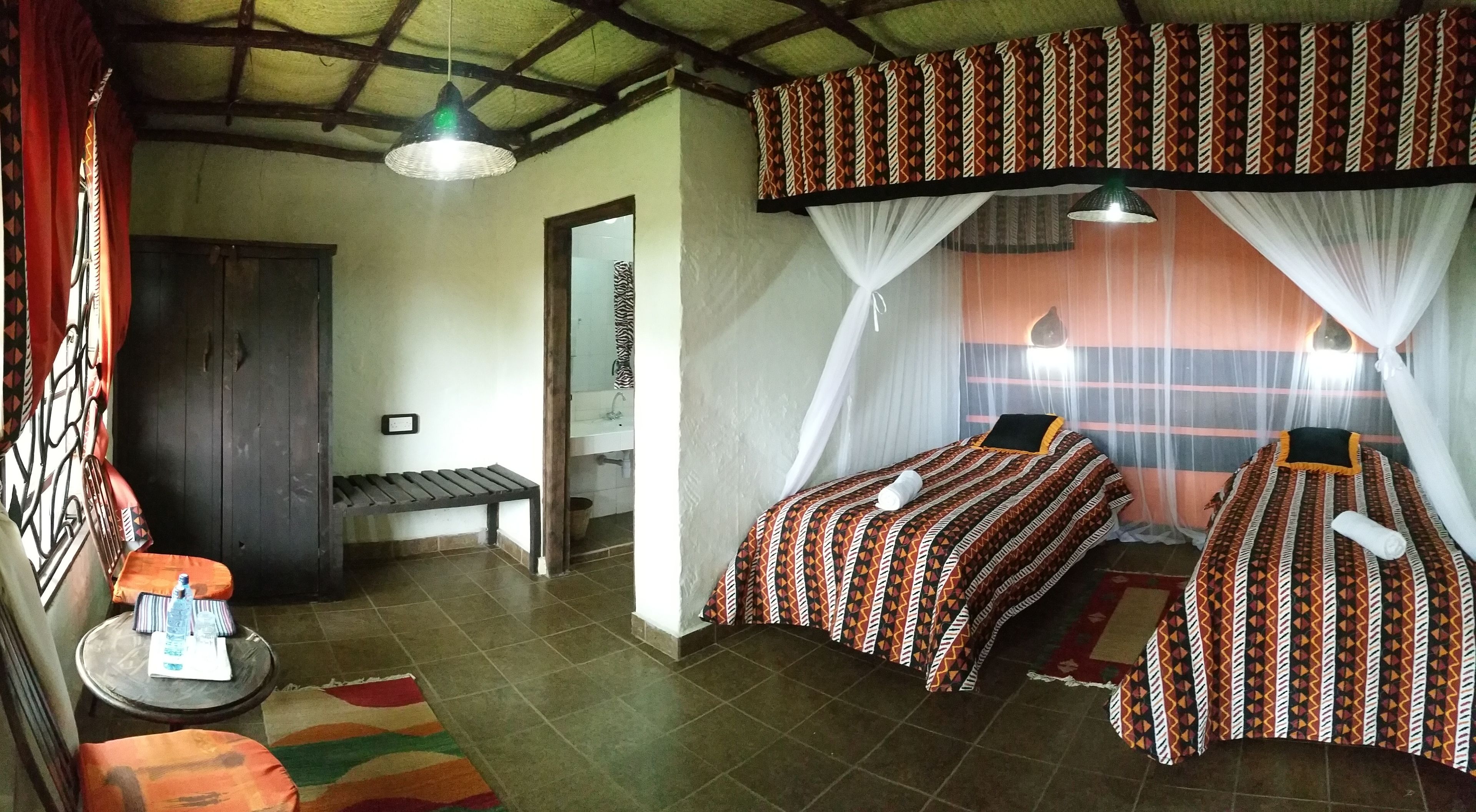A room with two beds, a bench, a small table with a water bottle, and a hanging light fixture.