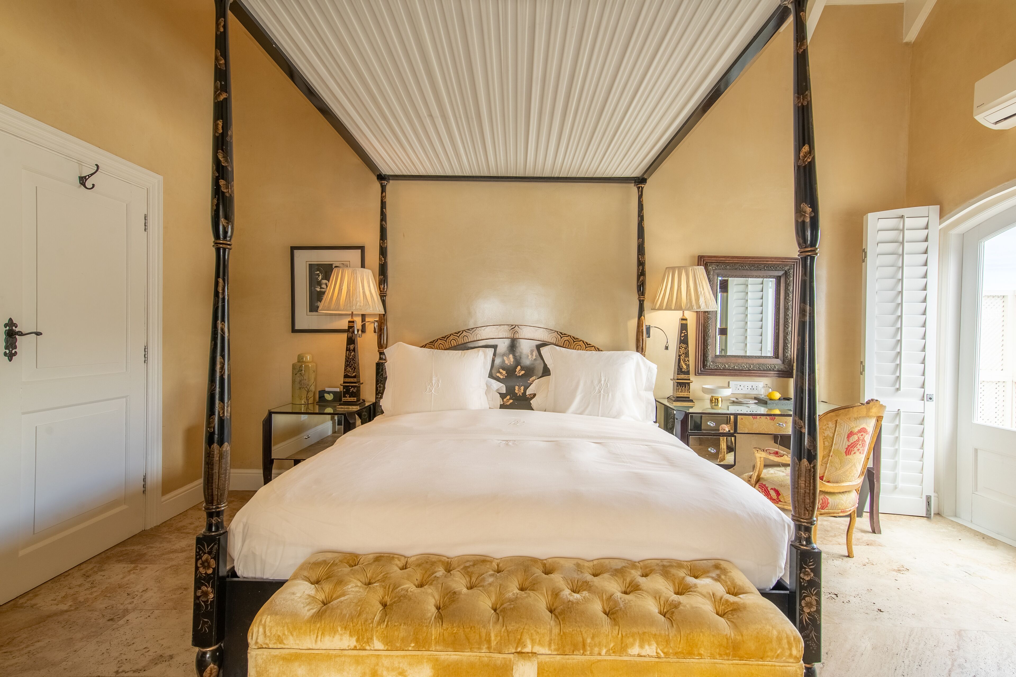 Una cama con dosel y cuatro columnas, con cabecera capitonada, una mesita de noche con lámpara y una silla en una habitación con pared color beige y techo blanco.