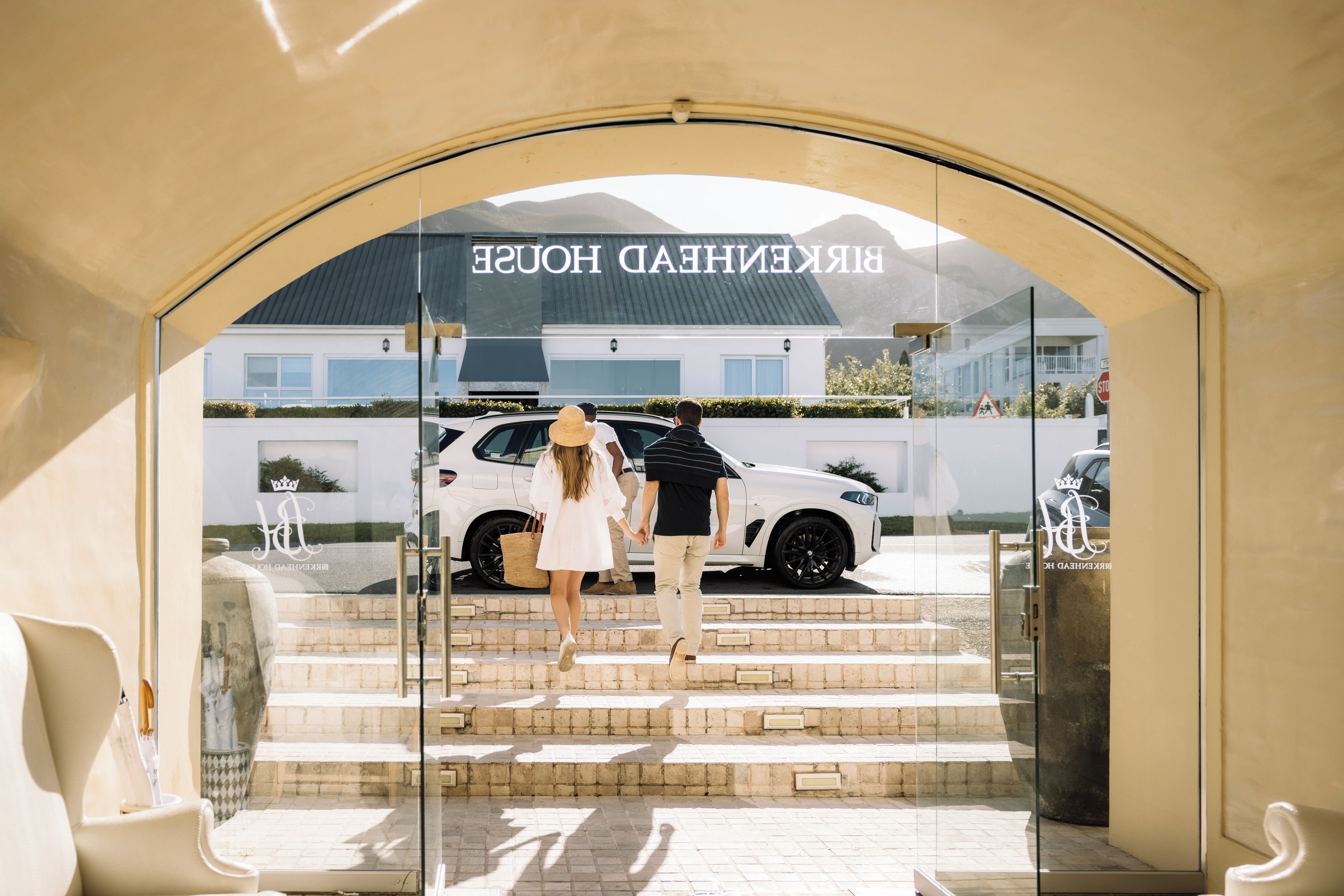 A hotel entrance with a glass facade, a white car parked outside, and a staircase leading to the entrance.