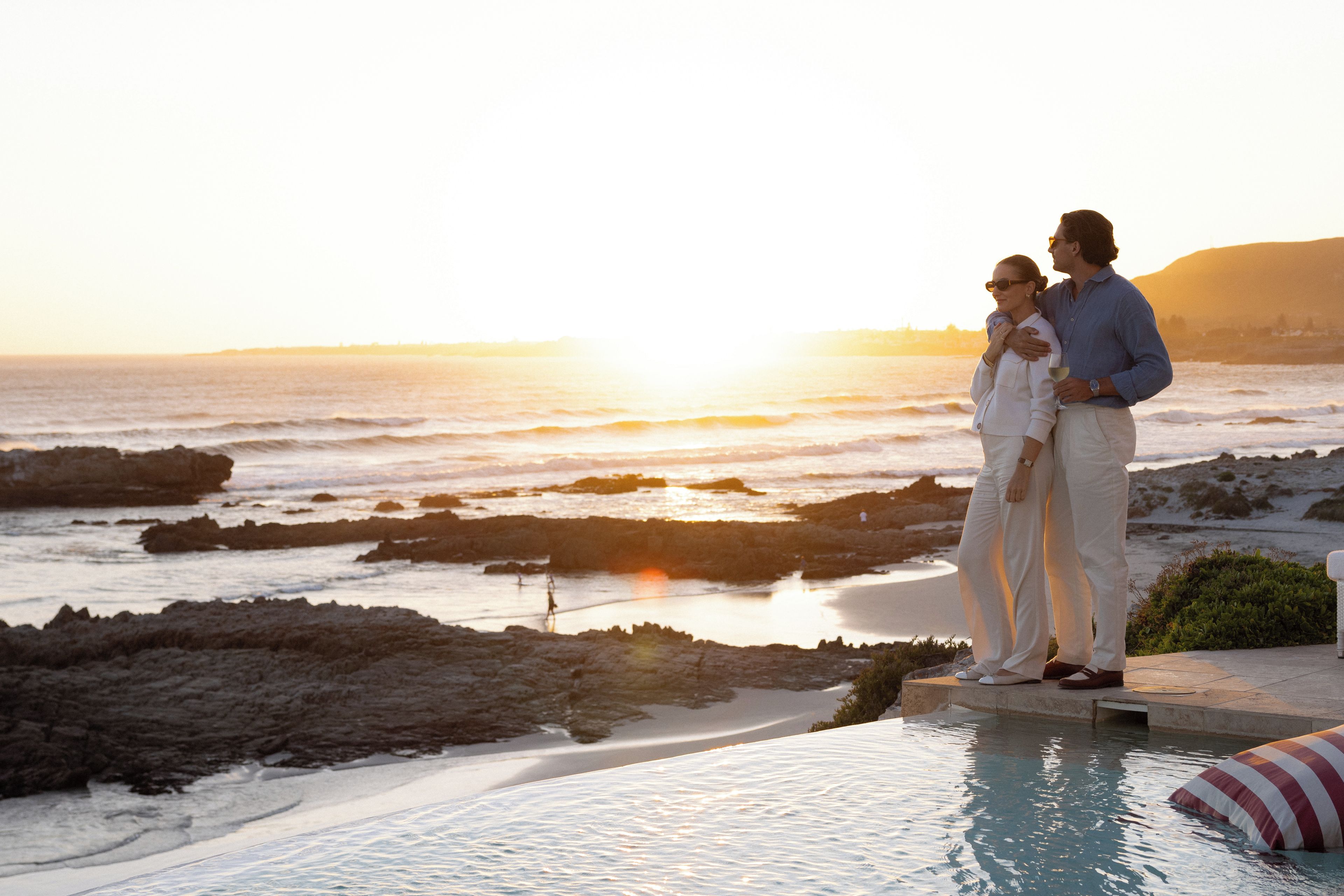 Una pareja parada junto a una piscina infinita con vistas a un litoral rocoso al atardecer.
