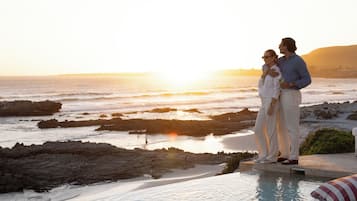 Una pareja parada junto a una piscina infinita con vistas a un litoral rocoso al atardecer.