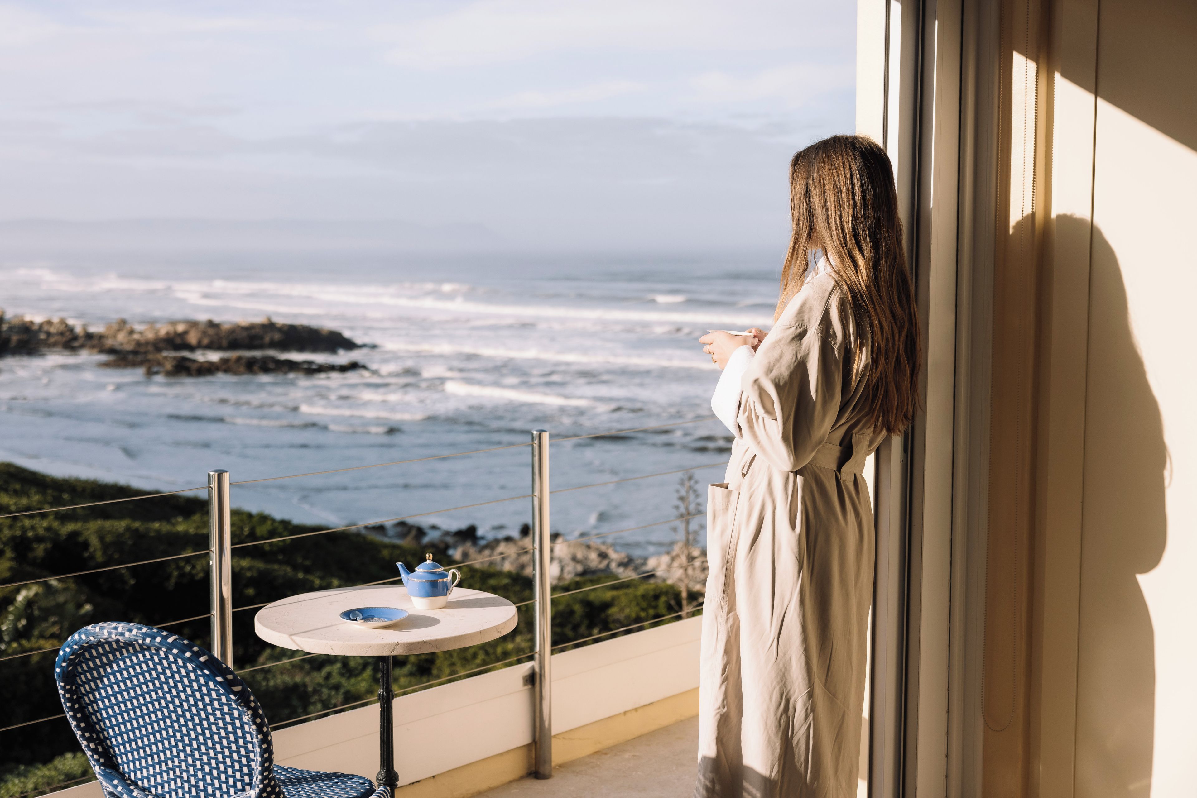 A balcony with a view of the ocean, a table with a teapot, and a chair.
