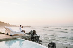 A man taking a photo with a camera, sitting on a lounge chair by a pool overlooking the ocean. - Birkenhead House (Hermanus)
