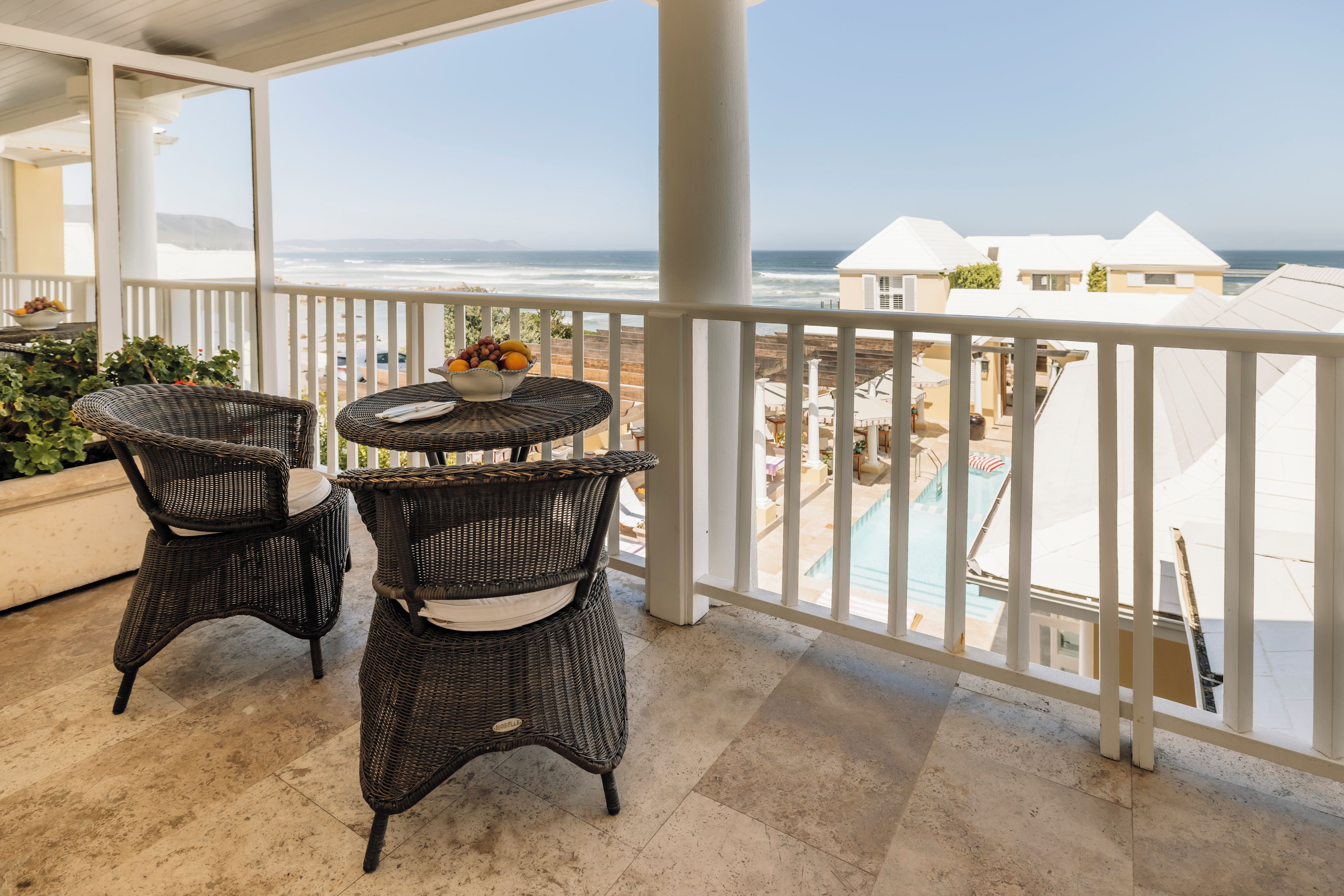 Ein Balkon mit Korbmöbeln und einem Tisch mit Früchten, mit Blick auf Strand und Poolbereich.