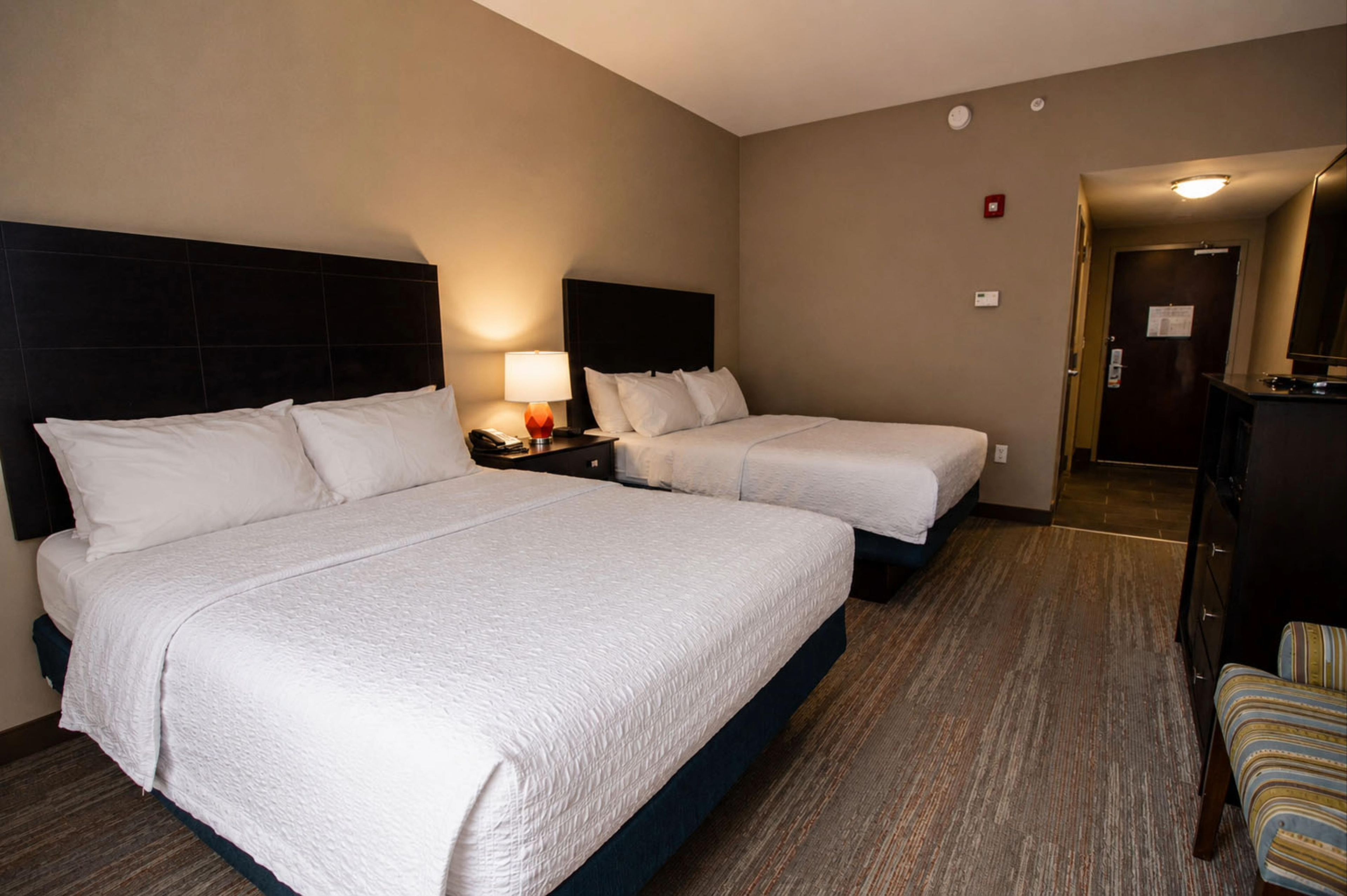 Une chambre d’hôtel avec deux lits, une table de chevet avec une lampe, un bureau avec un ordinateur et une porte donnant sur une autre pièce.