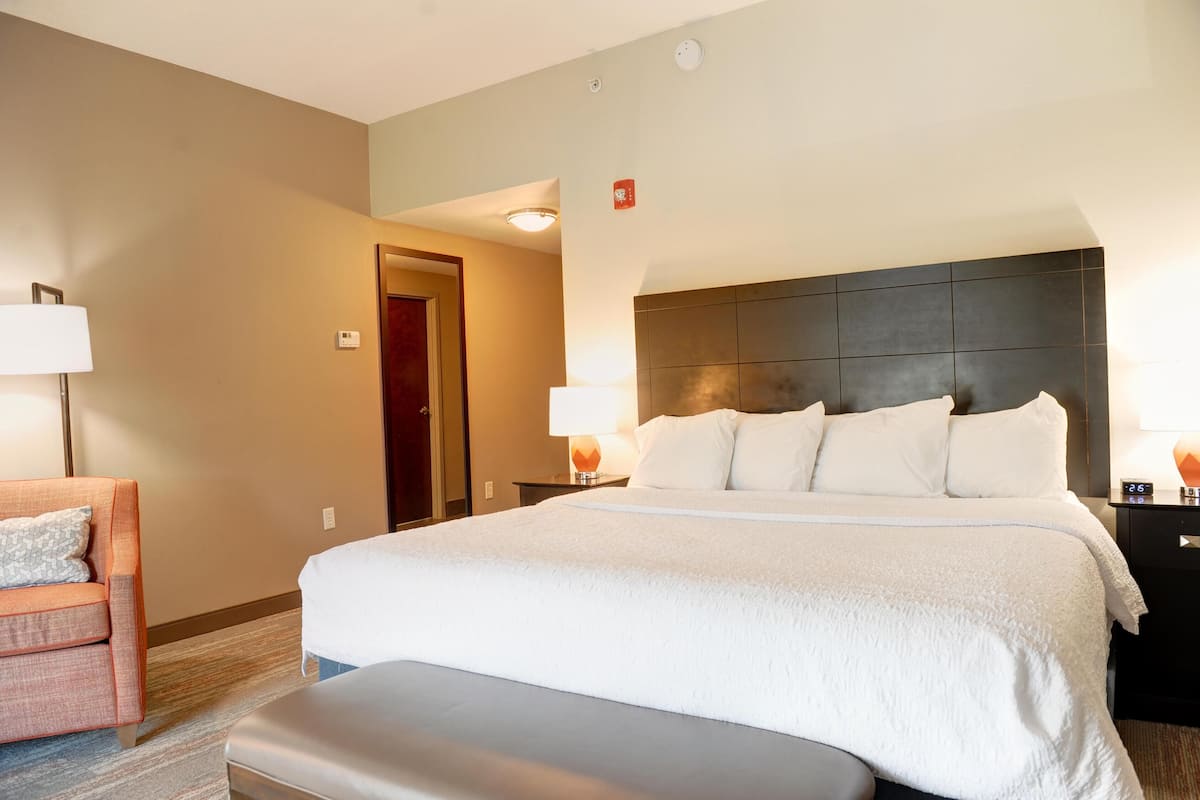 Quarto de hotel com uma cama grande, uma mesinha de cabeceira com abajur, uma cadeira e uma pequena mesa com relógio.