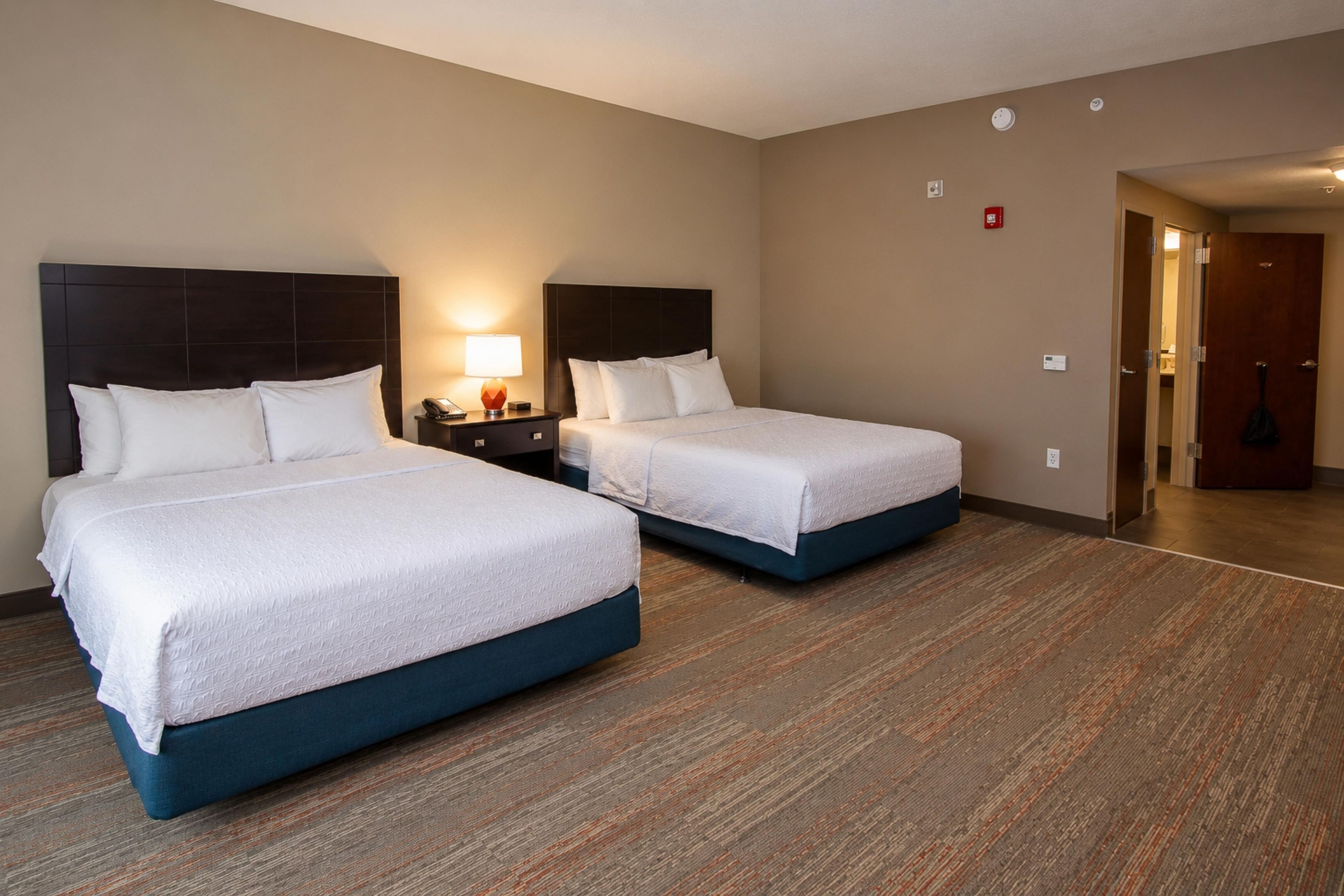 Habitación de hotel con dos camas, una mesita de noche con lámpara y acceso a un baño.