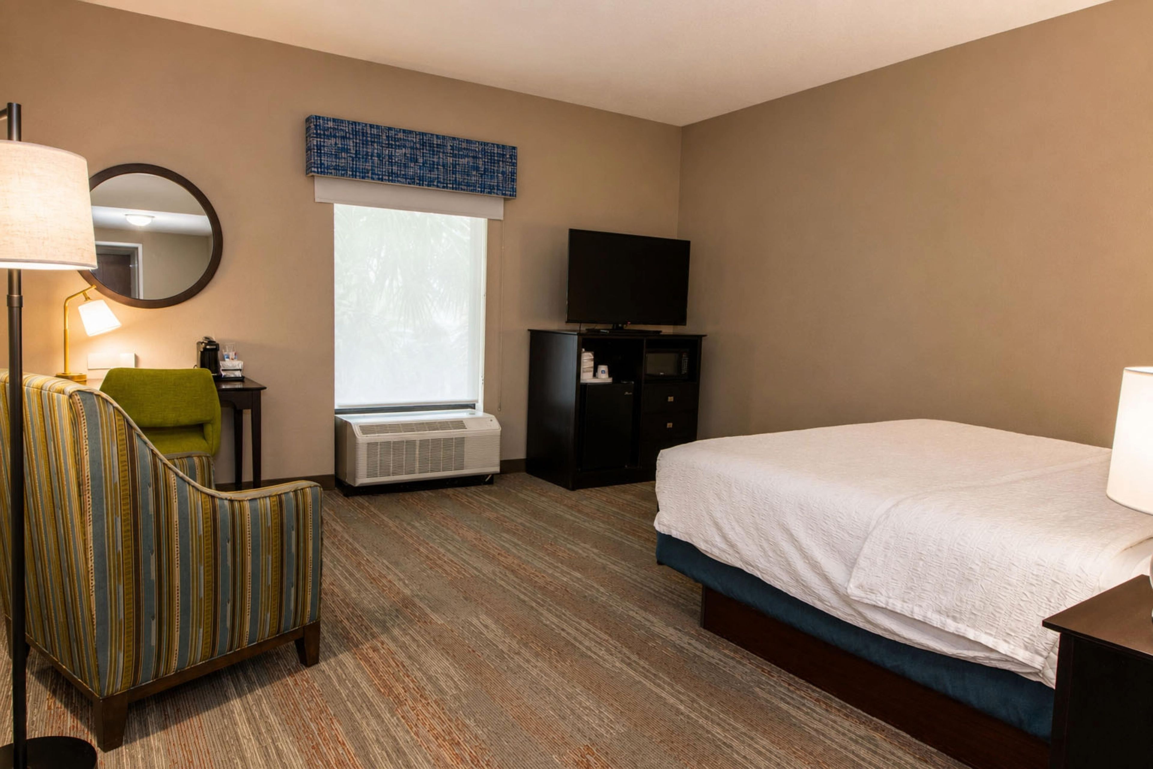 A hotel room with a bed, a chair, a desk with a lamp, a television, and a window.