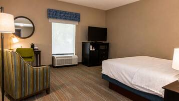 A hotel room with a bed, a chair, a desk with a lamp, a television, and a window.