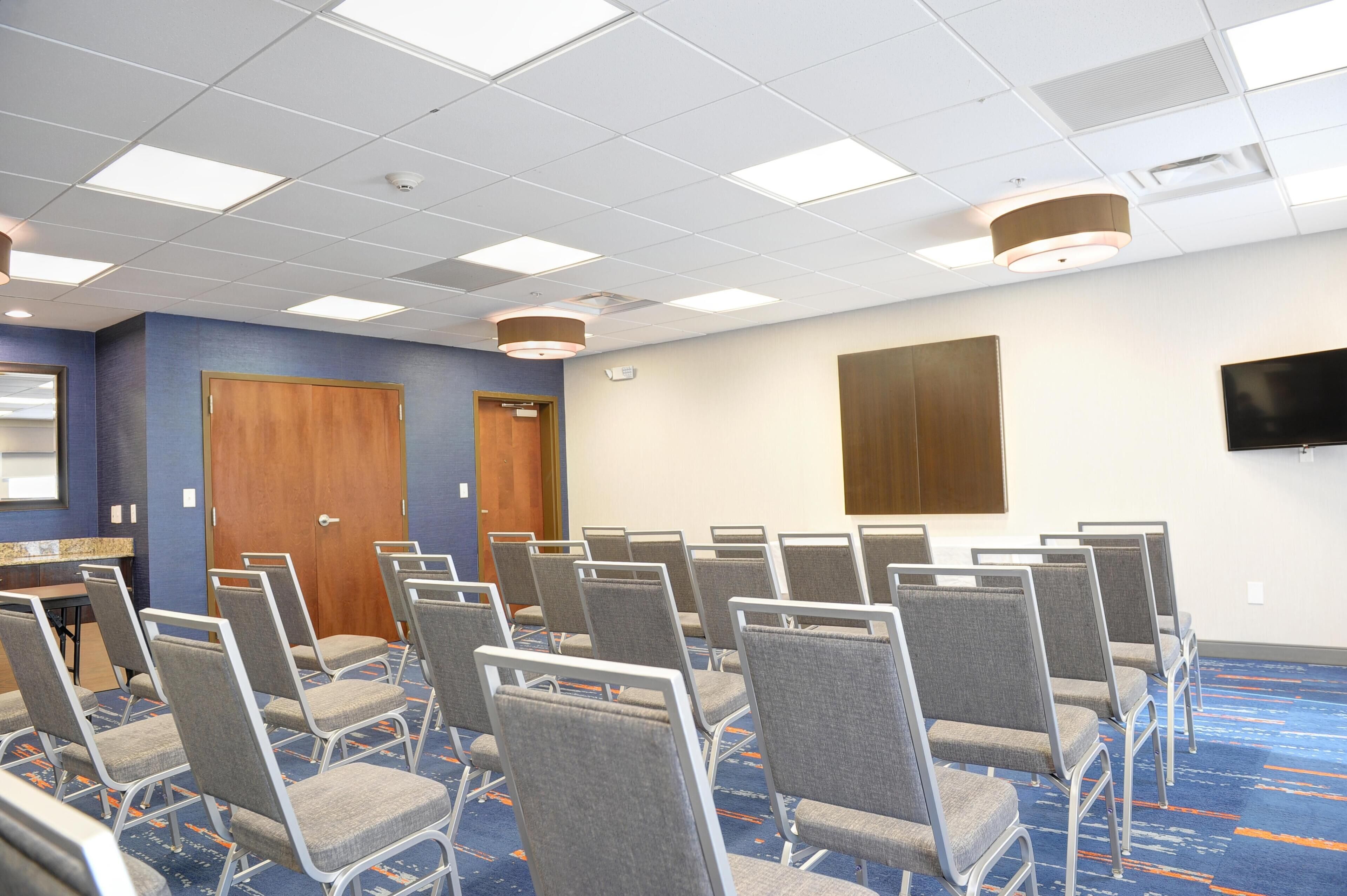 Un salón de conferencias con filas de sillas, una puerta de madera, un televisor en la pared y un techo con luces cuadradas.