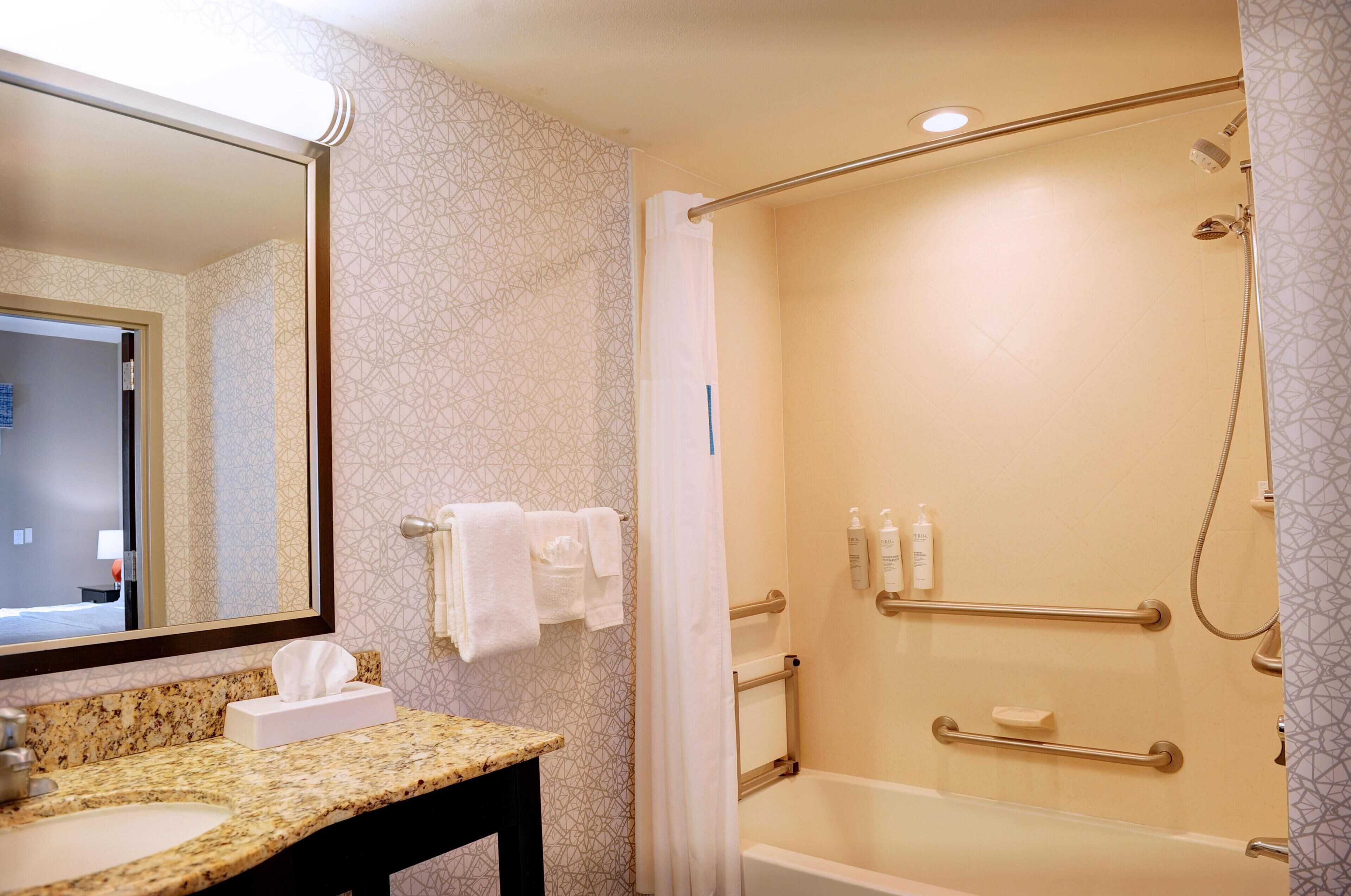 A bathroom with a sink, mirror, shower, and handrails.