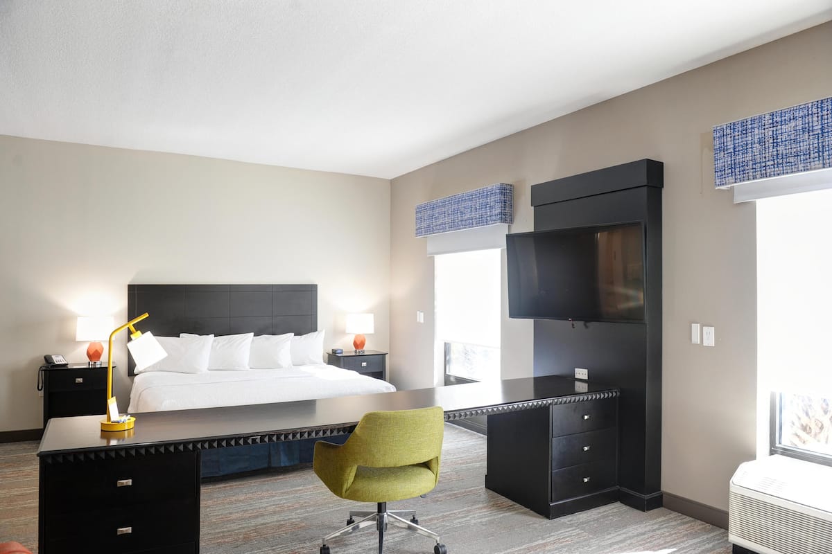 Quarto de hotel com cama, escrivaninha, cadeira, televisão e janela com persianas.