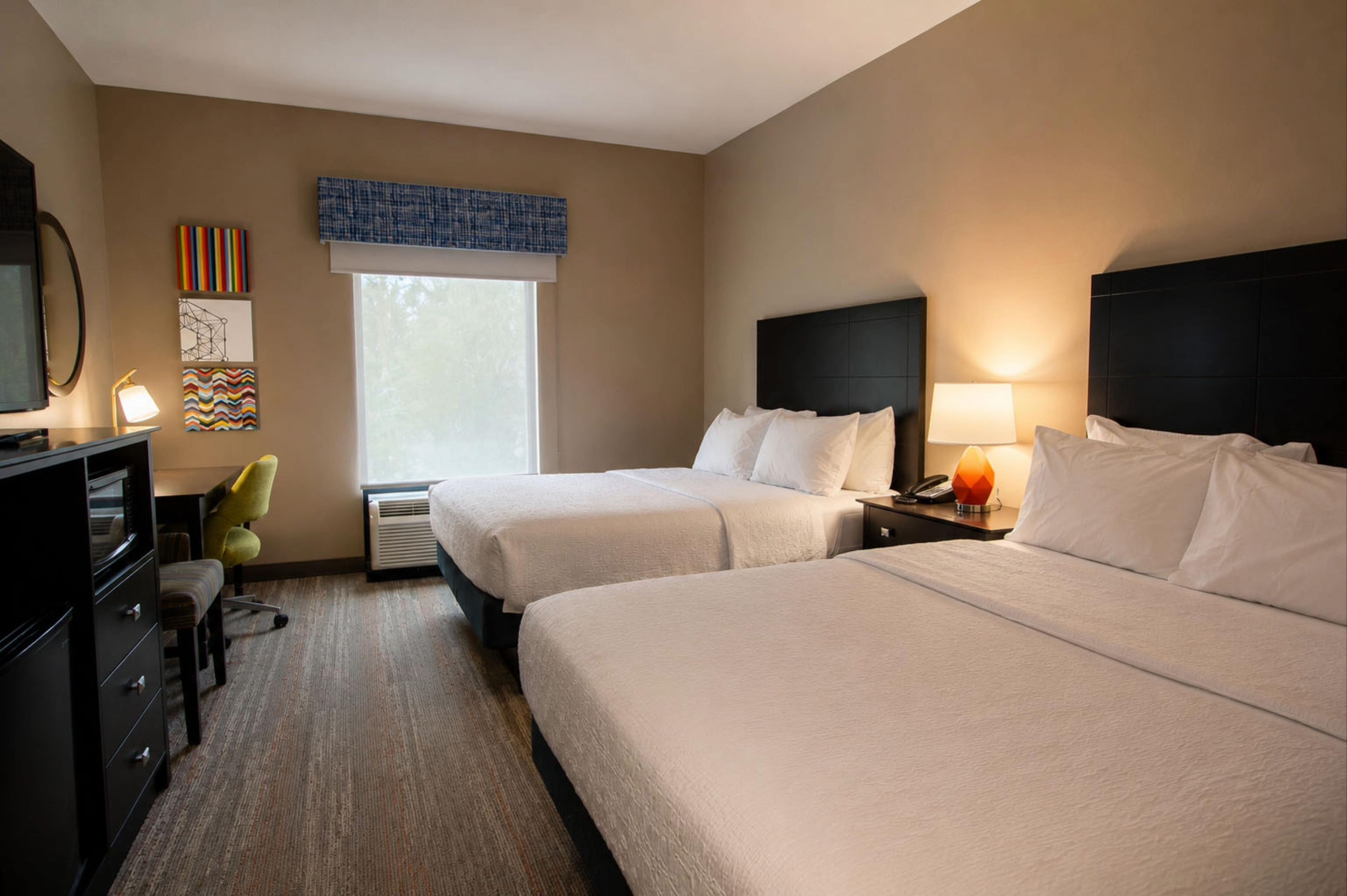 Habitación de hotel con dos camas, un escritorio con lámpara, una silla y una ventana con persianas.