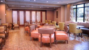 A hotel dining area with leather seating, a mix of round and rectangular tables, and large windows with curtains.