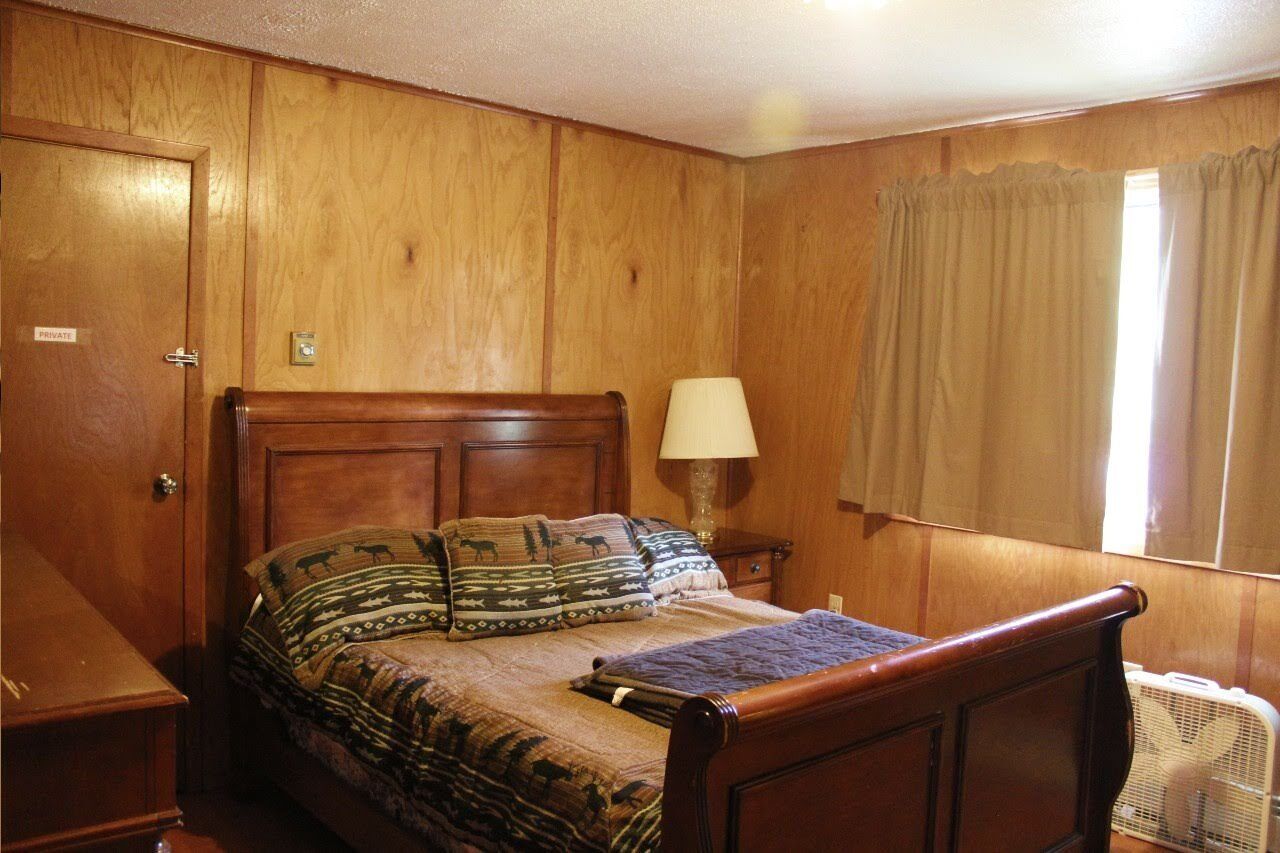Ein Zimmer mit Holzwänden, einem Bett, einer Nachttischlampe und einem Fenster mit Vorhängen.