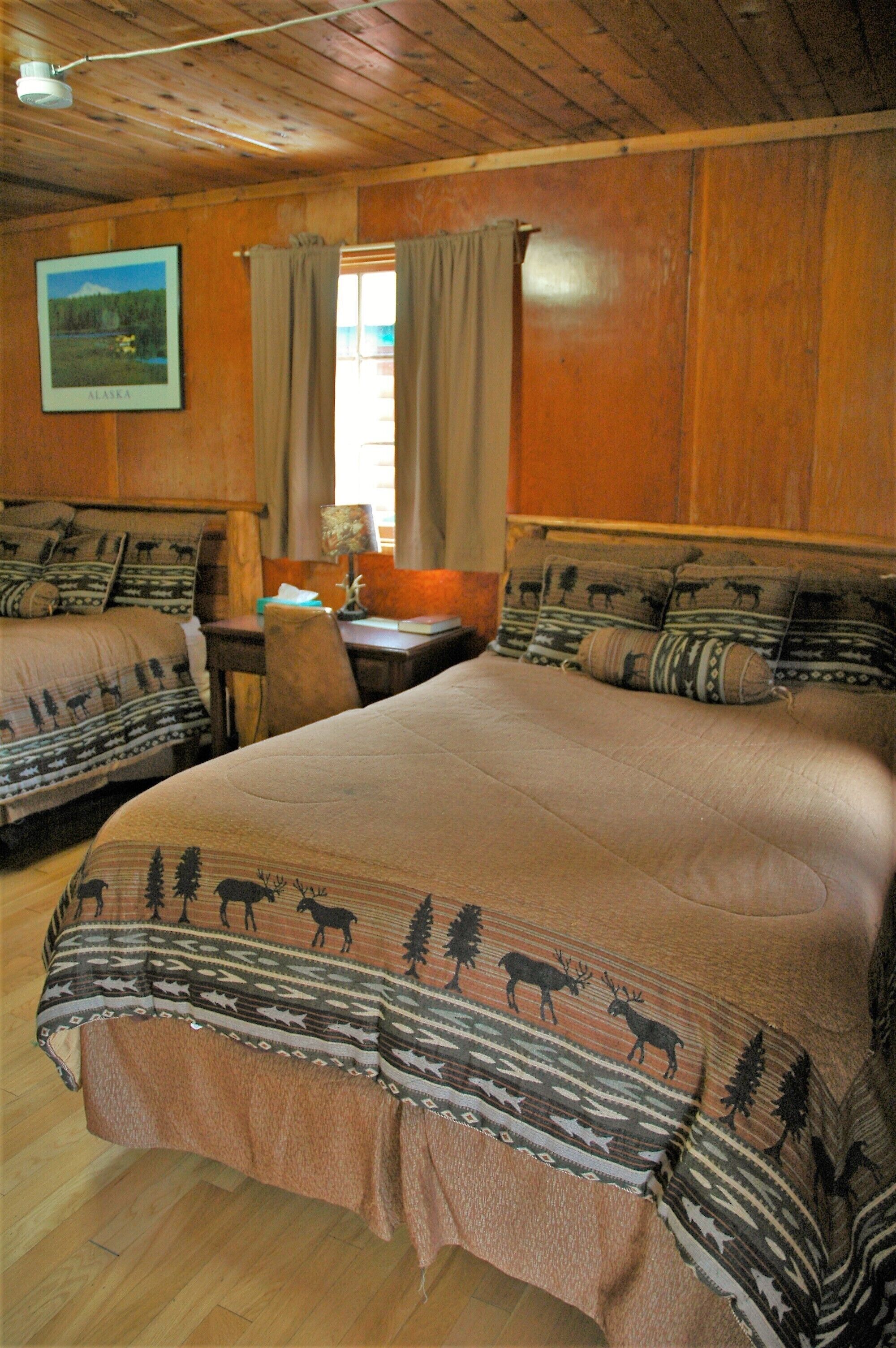 A room with two beds, wooden walls, a framed picture of Alaska, and a window with curtains.