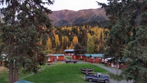 Una cabaña con chozas, una camioneta y un bote en un valle boscoso con montañas al fondo.