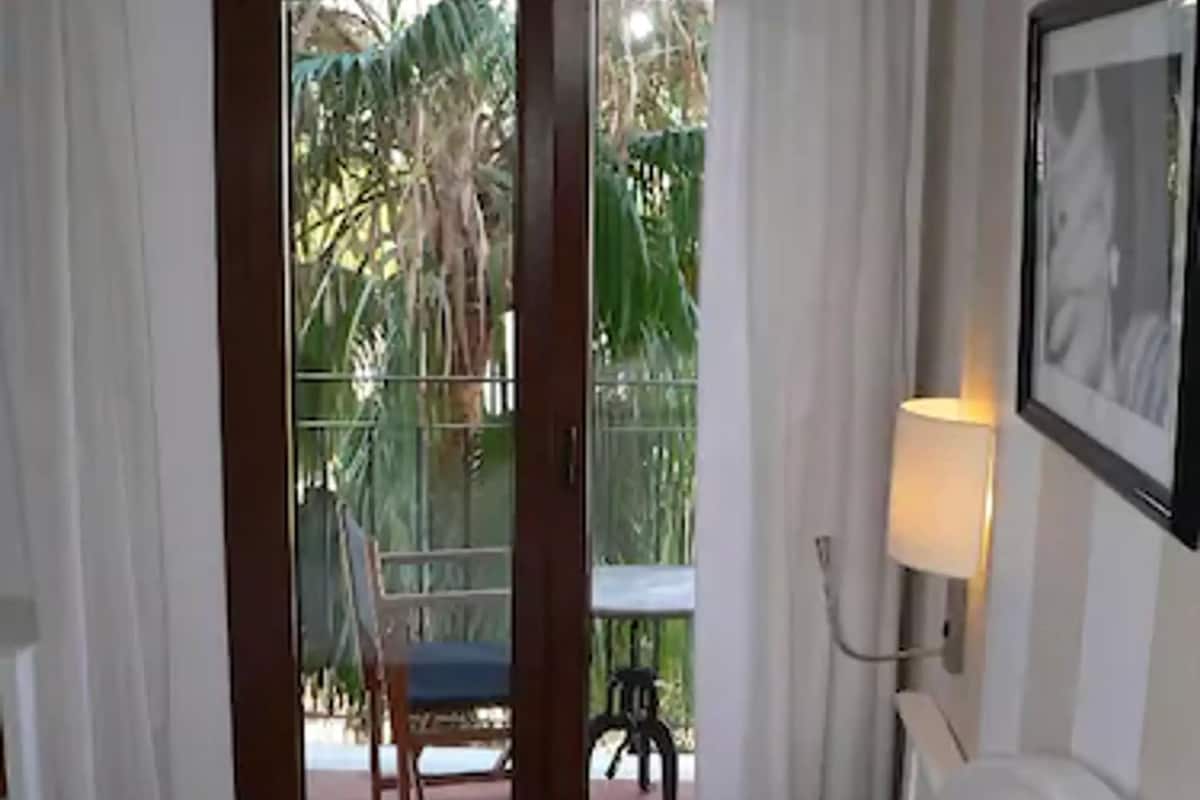 A hotel room with a bed, a chair, a table, and a balcony with a view of palm trees.