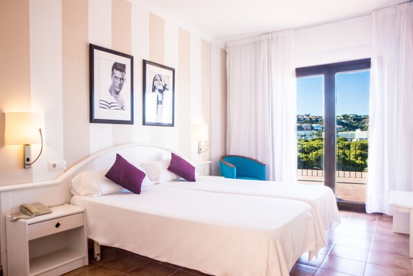 Habitación de hotel con dos camas, una mesita de noche con teléfono, una silla y un balcón con vistas.