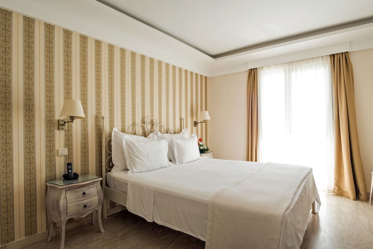 Une chambre d’hôtel avec un grand lit, deux tables de chevet, une petite table, une lampe et une fenêtre avec des rideaux.