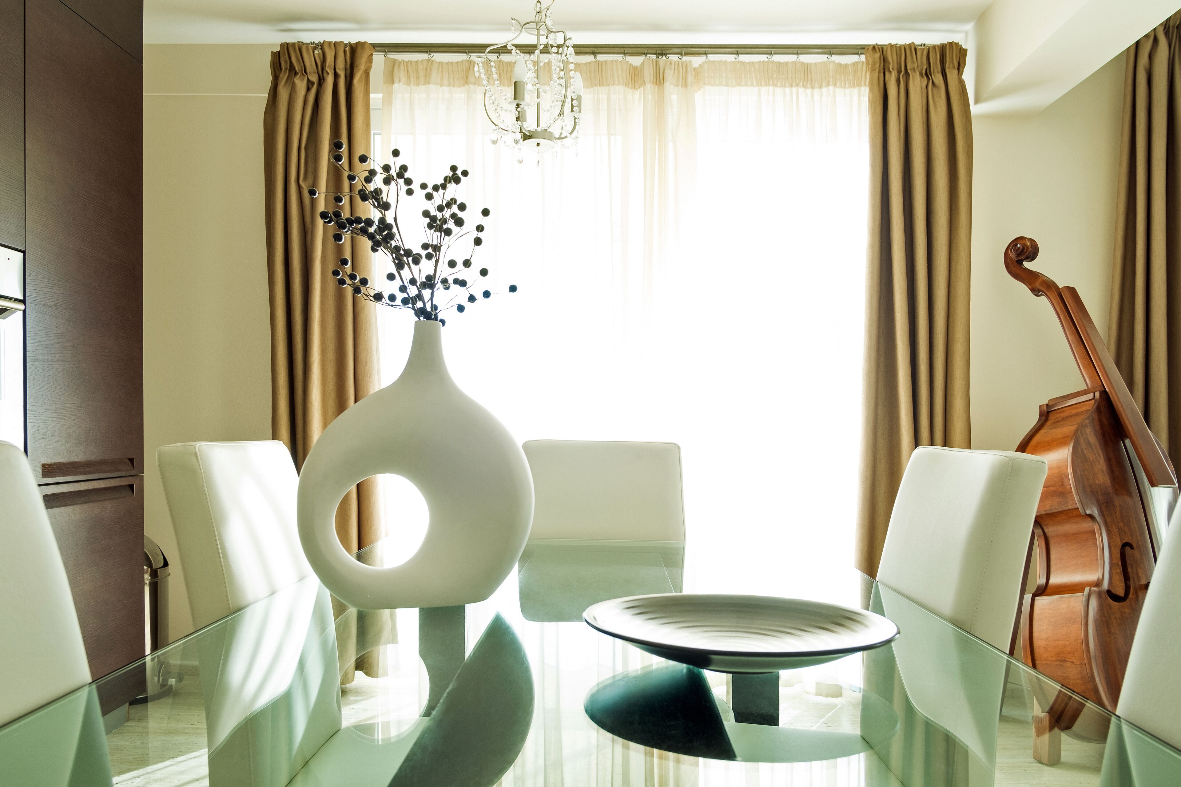 A dining area with a glass table, white chairs, a vase with flowers, and a cello in the background.